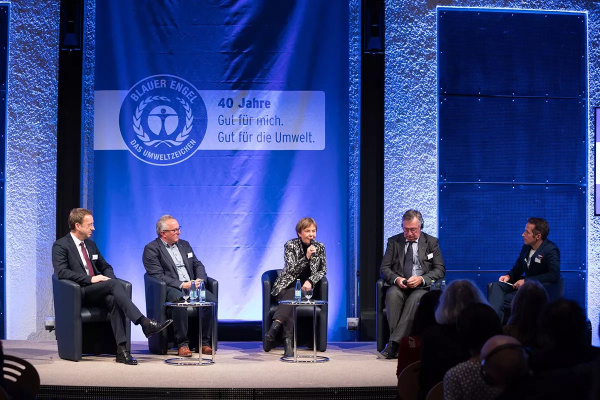 Four men in suits sit on stage, in the middle a woman with a microphone