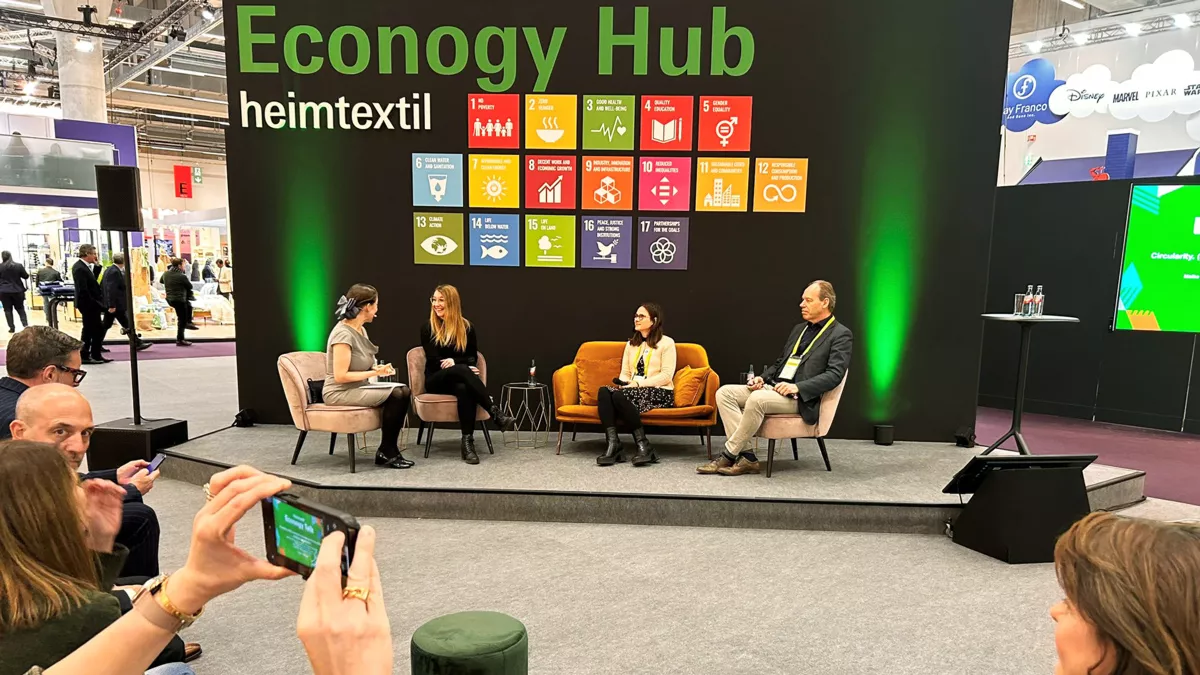 Three women and one man in conversation on a podium with an audience. “Econogy Hub, heimtextil” is written on a partition wall