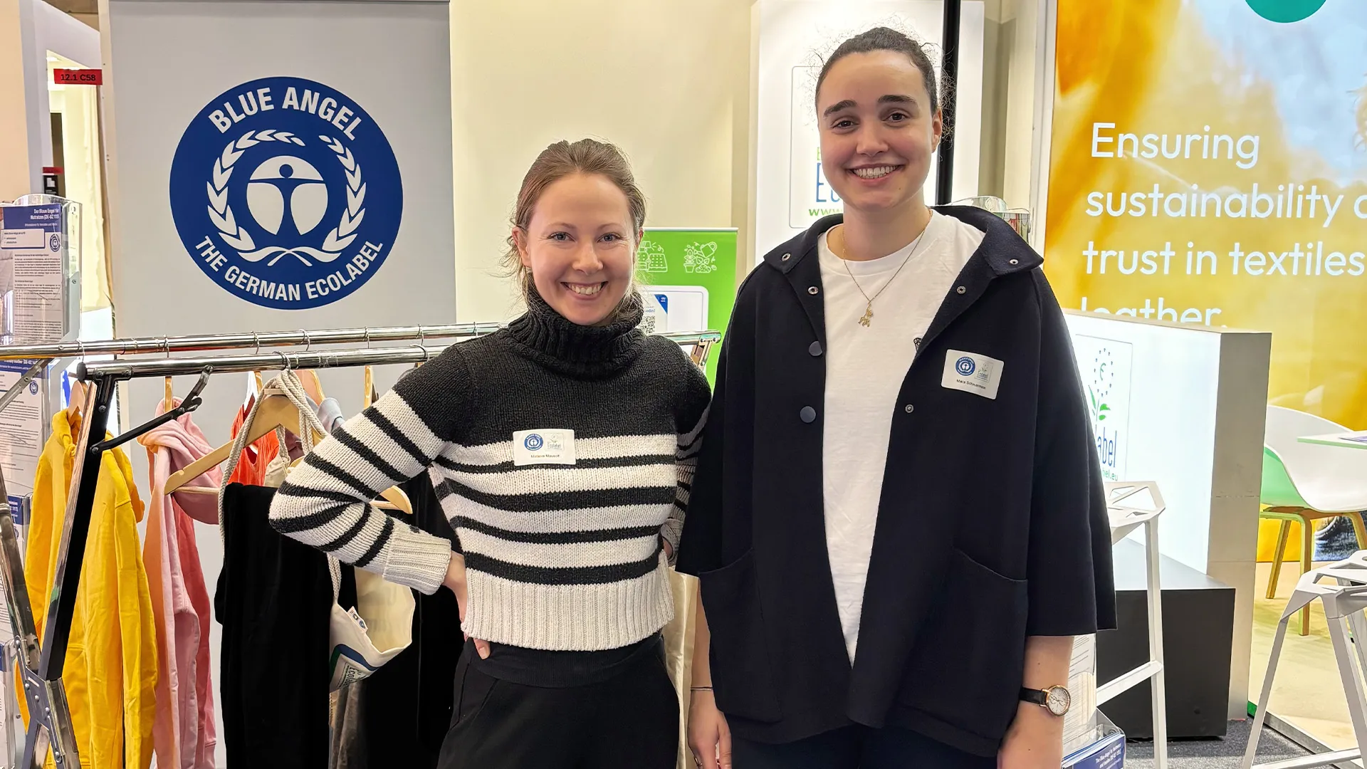 Two smiling women stand in front of the Blue Angel logo and a clothes rack