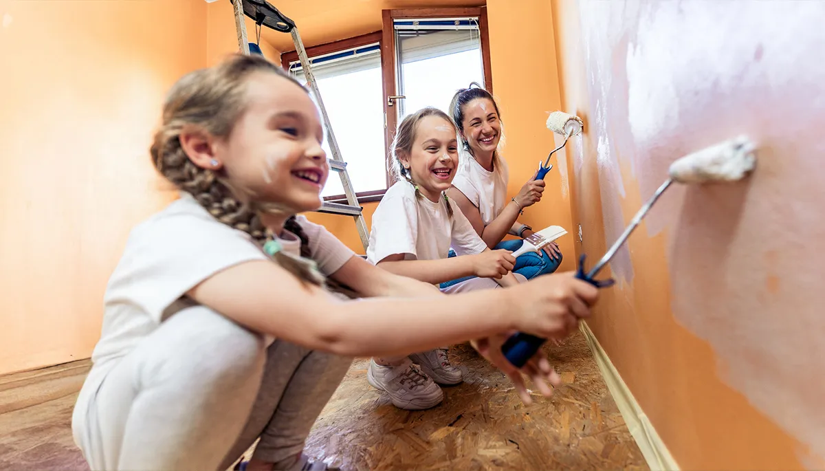 Two girls and a woman laugh as they paint a wall.