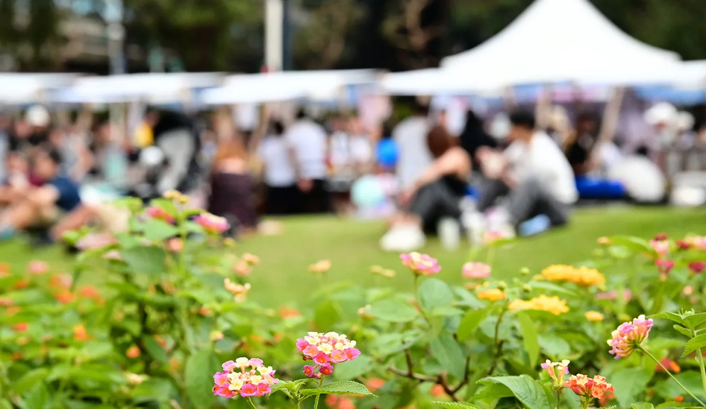 Unscharf im Hintergrund sieht man 3-4 Zelte einer Outdoor-Veranstaltung und viele Personen die drumrum stehen. Im Vordergrund sind Blumen.
