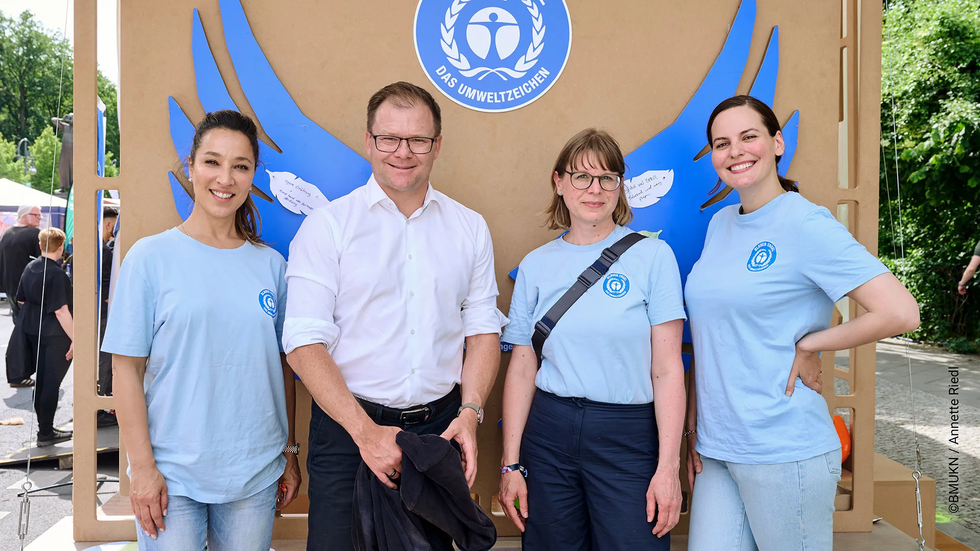Photo of 4 people: Federal Environment Minister Carsten Schneider visits the Blue Angel stand, in the background a brown wall with the Blue Angel logo and blue wings.
